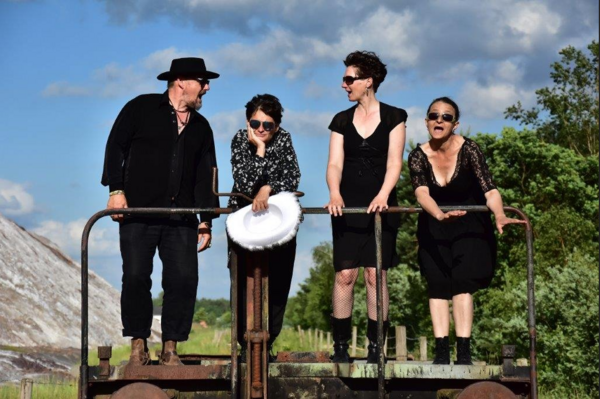 Vier Personen (drei Frauen, ein Mann) stehen hinter einem Geländer und schauen sich an oder herunter. Sie sind schwarz gekleidet.