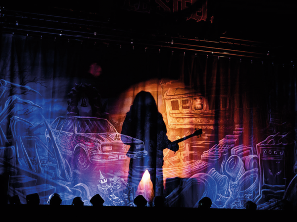 Eine schwarz-bunte, gezeichnete Bühnenkulisse auf Leinwand. Mittig der Schatten einer langhaarigen Person mit Gitarre.