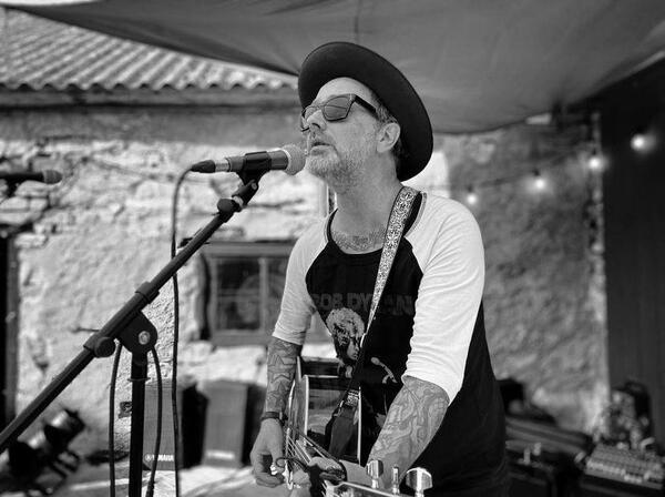 Ein Man mit Tattoos, schwarzem Hut, schwarzer Sonnenbrille und akustischer Gitarre singt in ein Mikrophon. Er ist leicht vornübergebeugt und es handelt sich um eine schwarz-weiß Aufnahme.