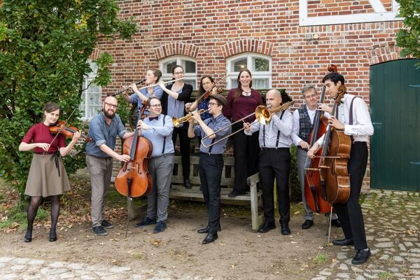 Elf Personen vor einem roten Backsteingebäude mit ihren klassischen Instrumenten. Sie spielen oder schauen lachend in die Luft.