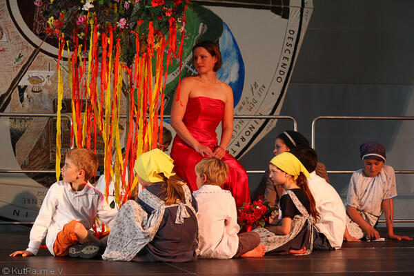 Eine Frau mit rotem Kleid sitzt auf einer Bühne mit Kindern in Kostümen.