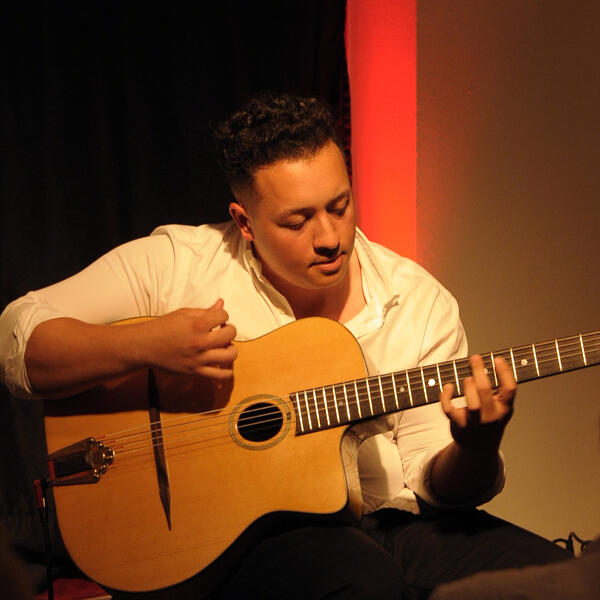 Ein junger Mann in weißem Oberteil und schwarzer Hose sitzt vor einem beleuchteten Hintergrund und spielt auf einer Akustikgitarre. Er schaut dabei auf seine Gitarre.