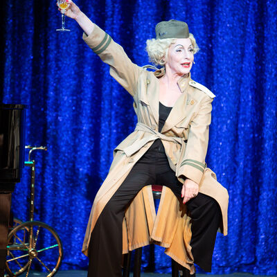 Eine Frau, Marlene Dietrich ähnlich, sitzt auf einem hohen Hocker und prostet an der Kamera vorbei mit einem Weißwein-Weinglas dem Publikum zu. 